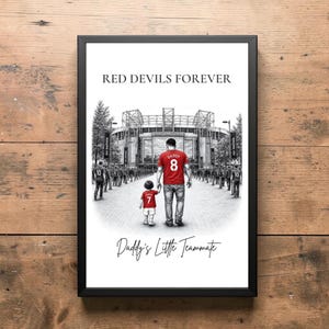 Op de afbeelding: Ingelijste print met een vader en kind in rode shirts met de nummers 8 en 7, lopend naar een stadion. De tekst "RED DEVILS FOREVER" en "Daddy's Little Teammate" zijn weergegeven.