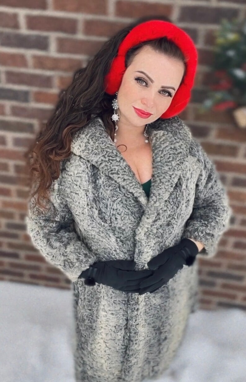 May include: A woman wearing a long, gray faux fur coat with a red earmuff headband. She is wearing black gloves and has long, dark hair.