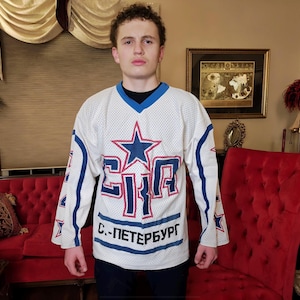 Peut inclure: Un chandail de hockey blanc avec des bordures bleues et rouges. Le chandail présente une grande étoile bleue avec un contour rouge et le texte "СКА" en rouge avec un contour bleu. Sous le texte se trouve "с. - ПЕТЕРБУРГ" en bleu. Le chandail est porté par une personne.