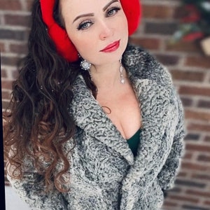 May include: A woman wearing a gray faux fur coat with a red earmuff. She is wearing a green top and silver jewelry.