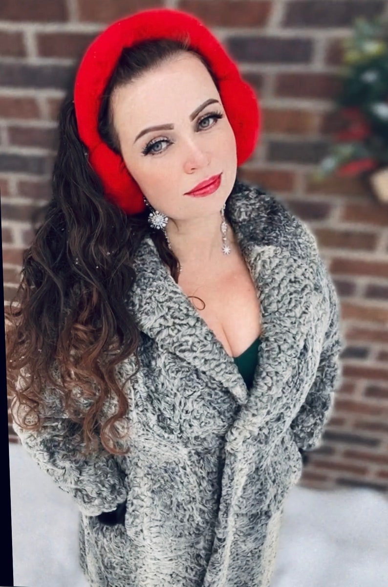 May include: A woman wearing a gray faux fur coat with a red earmuff. She is wearing a green top and silver jewelry.