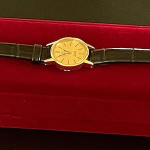 May include: A gold-tone wristwatch with a black leather strap on a red velvet watch box.