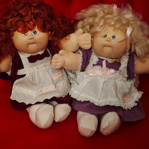 May include: Two Cabbage Patch Kids dolls, one with red hair and a purple dress, the other with blonde hair and a purple dress with white lace trim.