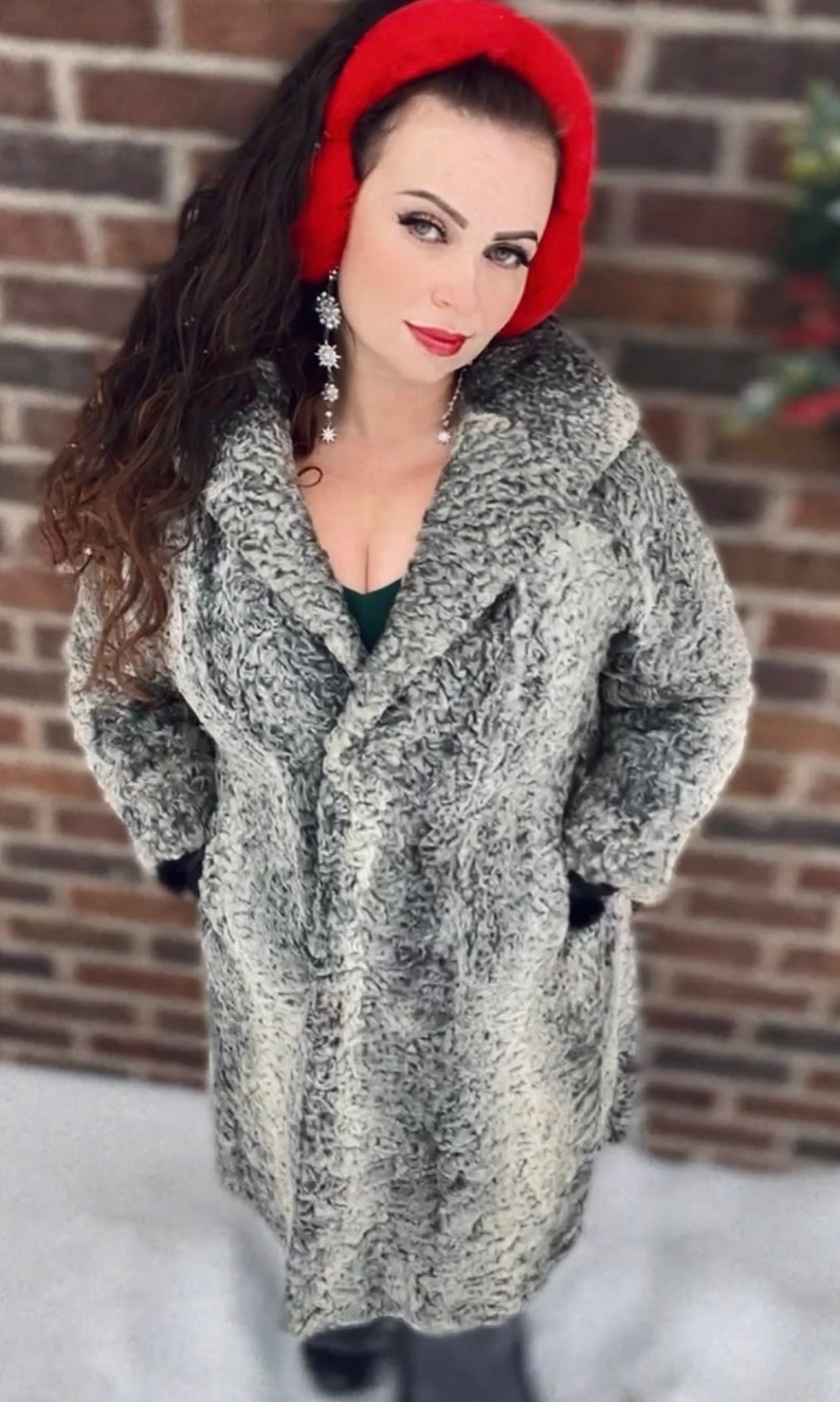 May include: A woman wearing a gray faux fur coat with a red ear warmer. She is standing in front of a brick wall.