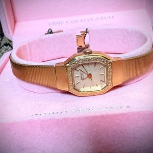 May include: A gold-toned, fine gold-plated wristwatch with a rectangular face and a textured band, presented in a pink velvet-lined box. The watch features a light-colored dial with gold hands and markers, and a decorative bezel.