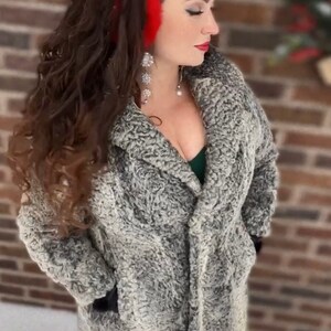 May include: A woman wearing a gray faux fur coat with a red headband. She is standing in front of a brick wall with snow on the ground.