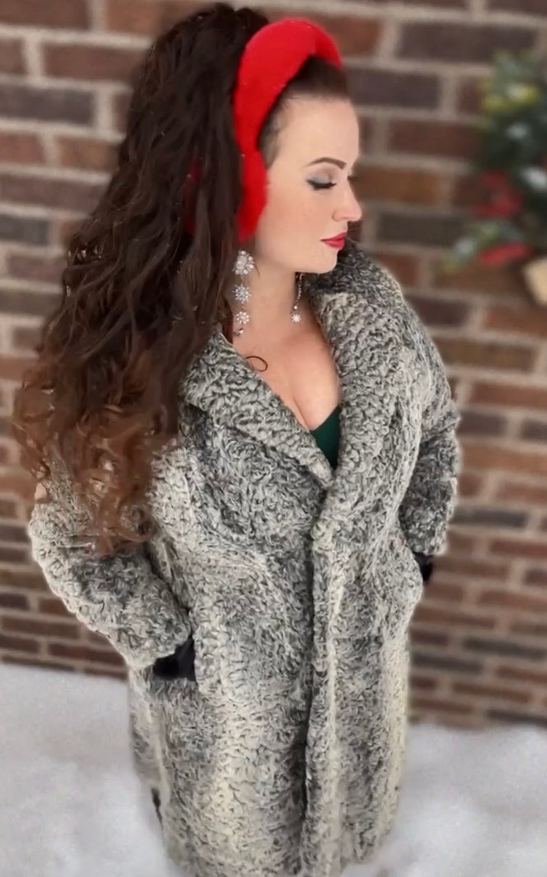 May include: A woman wearing a gray faux fur coat with a red headband. She is standing in front of a brick wall with snow on the ground.
