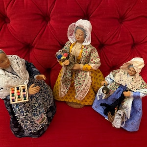 May include: Three antique figurines of women in traditional attire, set against a red backdrop. Each figure is dressed in a bonnet, shawl, and patterned dress, holding items like thread spools and a basket.
