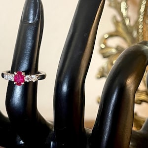 May include: A silver ring with a red oval gemstone surrounded by smaller white gemstones.