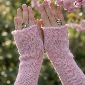Puede incluir: Un par de calentadores de brazo sin dedos de color rosa claro. Las mangas de punto son de un material suave y difuso. Los puños están ligeramente rizados. El fondo está borroso, con flores rosas y follaje verde.