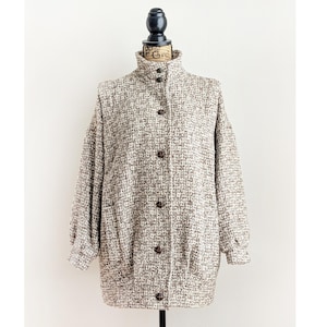 Op de afbeelding: Een tweed jasje met een hoge kraag en knoopsluiting. De jas is een mix van bruine en crèmekleuren, met een getextureerde weefsel. Het heeft lange mouwen en twee voorzakken. De jas wordt op een mannequin getoond.
