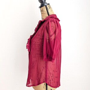 Op de afbeelding: Een bordeauxrode, semi-transparante blouse met korte mouwen en een gerimpelde kraag. De blouse wordt op een mannequin getoond en heeft een delicate, luchtige uitstraling. De stof lijkt een subtiel patroon te hebben. Een stijlvolle toevoeging aan elke garderobe.