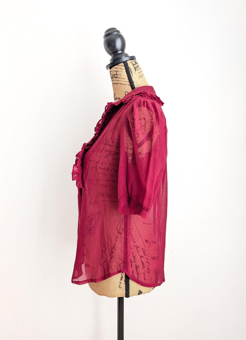 Op de afbeelding: Een bordeauxrode, semi-transparante blouse met korte mouwen en een gerimpelde kraag. De blouse wordt op een mannequin getoond en heeft een delicate, luchtige uitstraling. De stof lijkt een subtiel patroon te hebben. Een stijlvolle toevoeging aan elke garderobe.