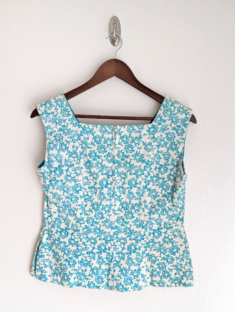 Pu&ograve; includere: Un top bianco senza maniche con un motivo floreale blu. Il top ha una scollatura quadrata e una chiusura a zip sul retro.