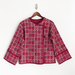May include: A quilted jacket in shades of red, burgundy, and cream, featuring a geometric pattern. The jacket has a round collar, long sleeves, and a button closure. The fabric appears to be a soft, lightweight material, suitable for layering.