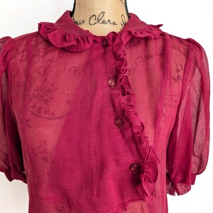 Op de afbeelding: Een doorschijnende, bordeauxrode blouse met een gerimpelde kraag en knoopsluiting. De top met korte mouwen heeft een delicate, semi-transparante uitstraling, met subtiele textuur zichtbaar in de stof. De blouse wordt op een mannequin getoond.