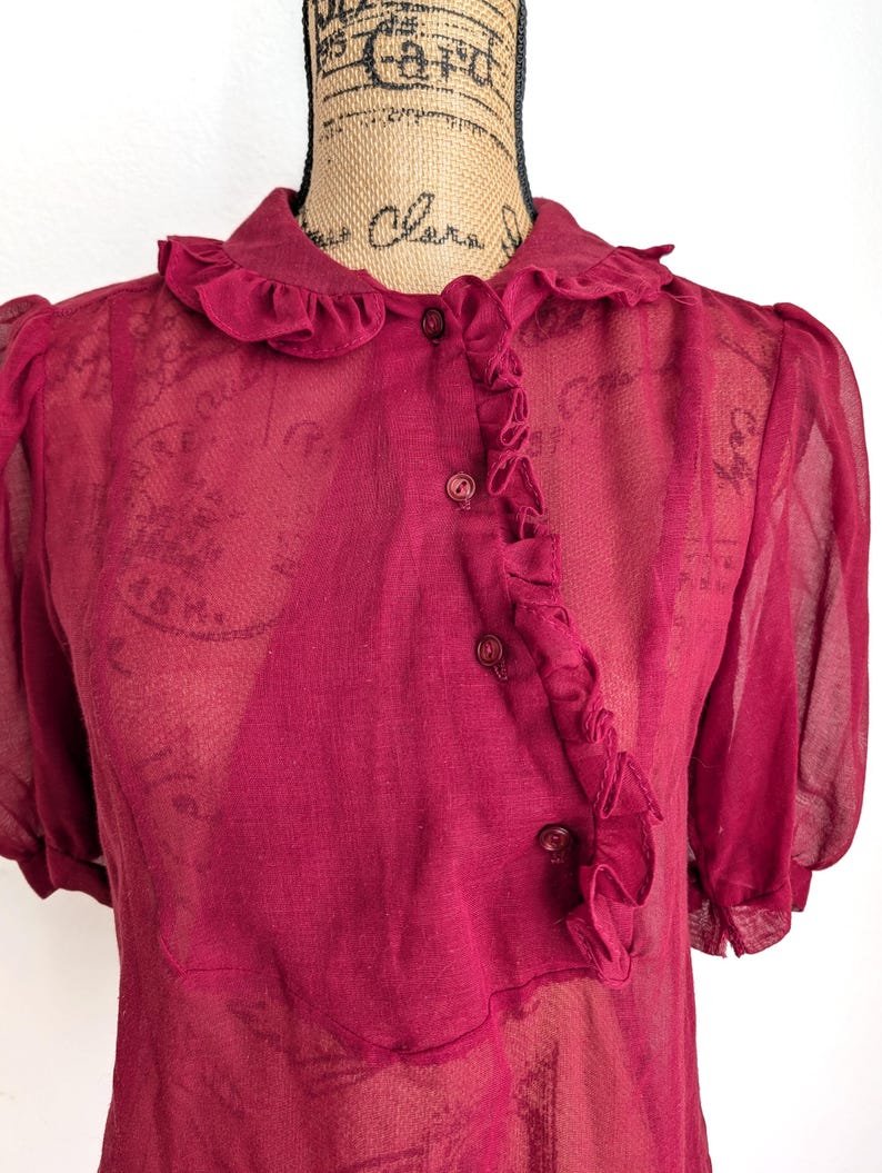 Op de afbeelding: Een doorschijnende, bordeauxrode blouse met een gerimpelde kraag en knoopsluiting. De top met korte mouwen heeft een delicate, semi-transparante uitstraling, met subtiele textuur zichtbaar in de stof. De blouse wordt op een mannequin getoond.