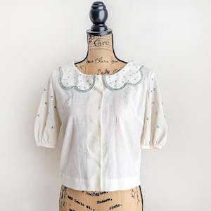 May include: Cream-colored blouse with a scalloped collar. The collar is adorned with small green embroidered details. The short sleeves have a slight puff, and the fabric has a subtle vertical texture. Small green floral embroidery accents the sleeves.