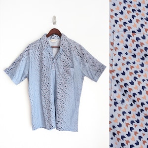 Puede incluir: Camisa azul claro de manga corta con cuello y bolsillo. La camisa presenta un patrón de pequeños puntos azul oscuro y rosa que se desvanecen hacia abajo. La camisa está colgada de una percha de madera.