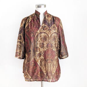 Peut inclure: Un chemisier à manches trois-quarts avec un col officier et une encolure en V. Le chemisier présente un motif cachemire dans des tons bordeaux, or et marron. Le tissu semble léger et légèrement transparent, avec un léger éclat.