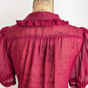 Op de afbeelding: Een doorschijnende, bordeauxrode blouse met korte, pofmouwen en een kraag met ruches. De achterkant van de blouse is zichtbaar en toont de delicate stof en het ontwerp. De blouse wordt op een mannequin gepresenteerd.