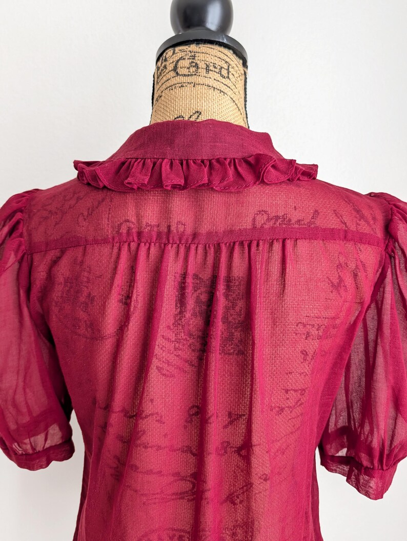 Op de afbeelding: Een doorschijnende, bordeauxrode blouse met korte, pofmouwen en een kraag met ruches. De achterkant van de blouse is zichtbaar en toont de delicate stof en het ontwerp. De blouse wordt op een mannequin gepresenteerd.