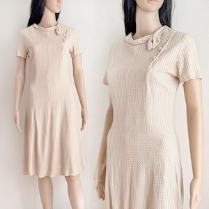 May include: A vintage beige dress with a textured pattern. The dress features short sleeves, a round collar, and decorative buttons. The dress has a fitted bodice and a flared skirt, creating a classic silhouette. The dress is displayed on a mannequin.