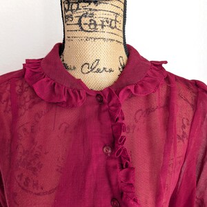 Op de afbeelding: Een bordeauxrode blouse met een gerimpelde kraag en knoopsluiting. De doorschijnende stof onthult een subtiel patroon. De top met korte mouwen wordt op een mannequin getoond.