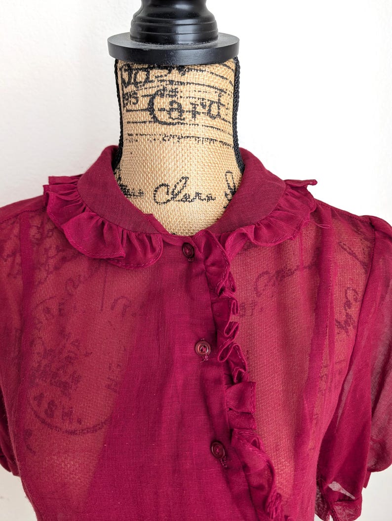 Op de afbeelding: Een bordeauxrode blouse met een gerimpelde kraag en knoopsluiting. De doorschijnende stof onthult een subtiel patroon. De top met korte mouwen wordt op een mannequin getoond.