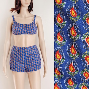 May include: A blue two-piece swimsuit featuring a paisley design in red, yellow, and white. The bikini top has thin straps, and the high-waisted shorts have white trim. The fabric is patterned with a repeating design.