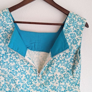 Pu&ograve; includere: Un abito senza maniche con sfondo bianco e motivo floreale blu. L'abito ha una fodera turchese e una cerniera sul retro.
