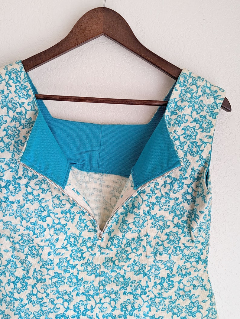 Pu&ograve; includere: Un abito senza maniche con sfondo bianco e motivo floreale blu. L'abito ha una fodera turchese e una cerniera sul retro.