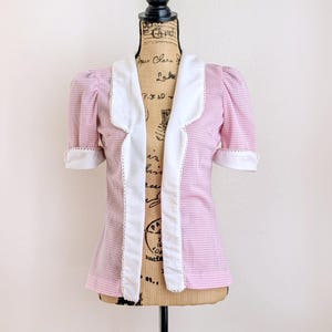 May include: Pink and white gingham short-sleeve top with a white collar and trim. The top has a fitted waist and puff sleeves. The white trim has a delicate lace detail. The top is displayed on a mannequin.