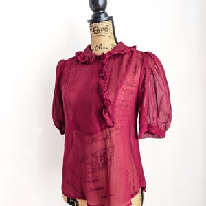 Op de afbeelding: Een bordeauxrode blouse met korte, gepofte mouwen en een gerimpelde kraag. De transparante stof drapeert over een mannequin en benadrukt de vintage stijl. De blouse heeft een knoopsluiting aan de voorkant en een delicate, romantische uitstraling.