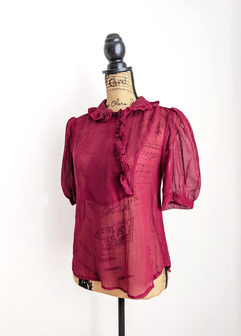 Op de afbeelding: Een bordeauxrode blouse met korte, gepofte mouwen en een gerimpelde kraag. De transparante stof drapeert over een mannequin en benadrukt de vintage stijl. De blouse heeft een knoopsluiting aan de voorkant en een delicate, romantische uitstraling.