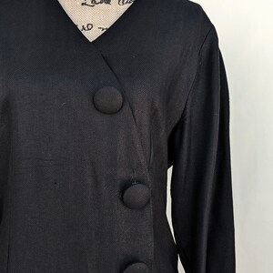 May include: A black dress with a V-neckline and a diagonal row of four large black buttons. The dress has a herringbone pattern and long sleeves.