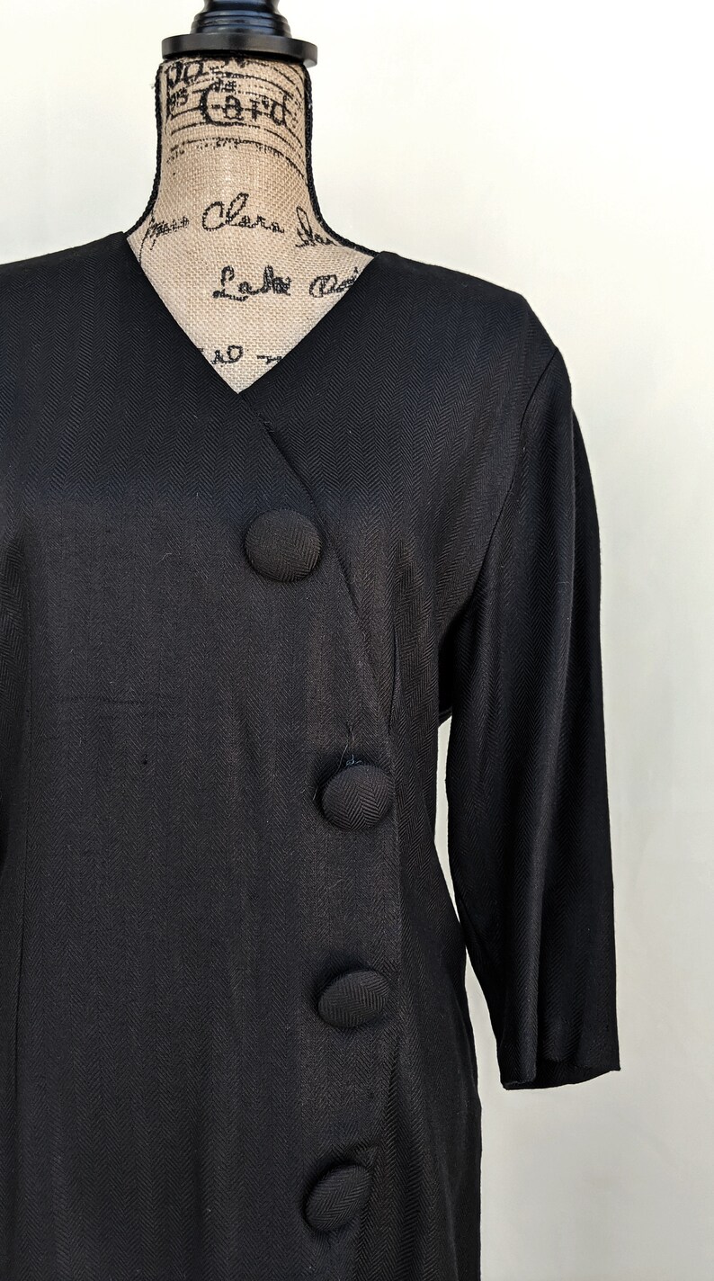 May include: A black dress with a V-neckline and a diagonal row of four large black buttons. The dress has a herringbone pattern and long sleeves.