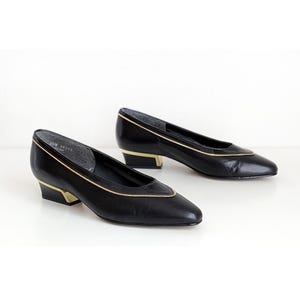 May include: A pair of black leather pumps with gold trim and a low, gold-colored heel. The shoes have a classic, pointed-toe design and are set against a white background. The shoes are a size 6.5M.