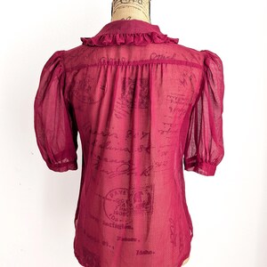 Op de afbeelding: Bordeaux transparante blouse met korte, gepofte mouwen en een kraag met ruches. De achterkant van de blouse is zichtbaar en toont een vintage printontwerp. De blouse wordt op een mannequin gepresenteerd.