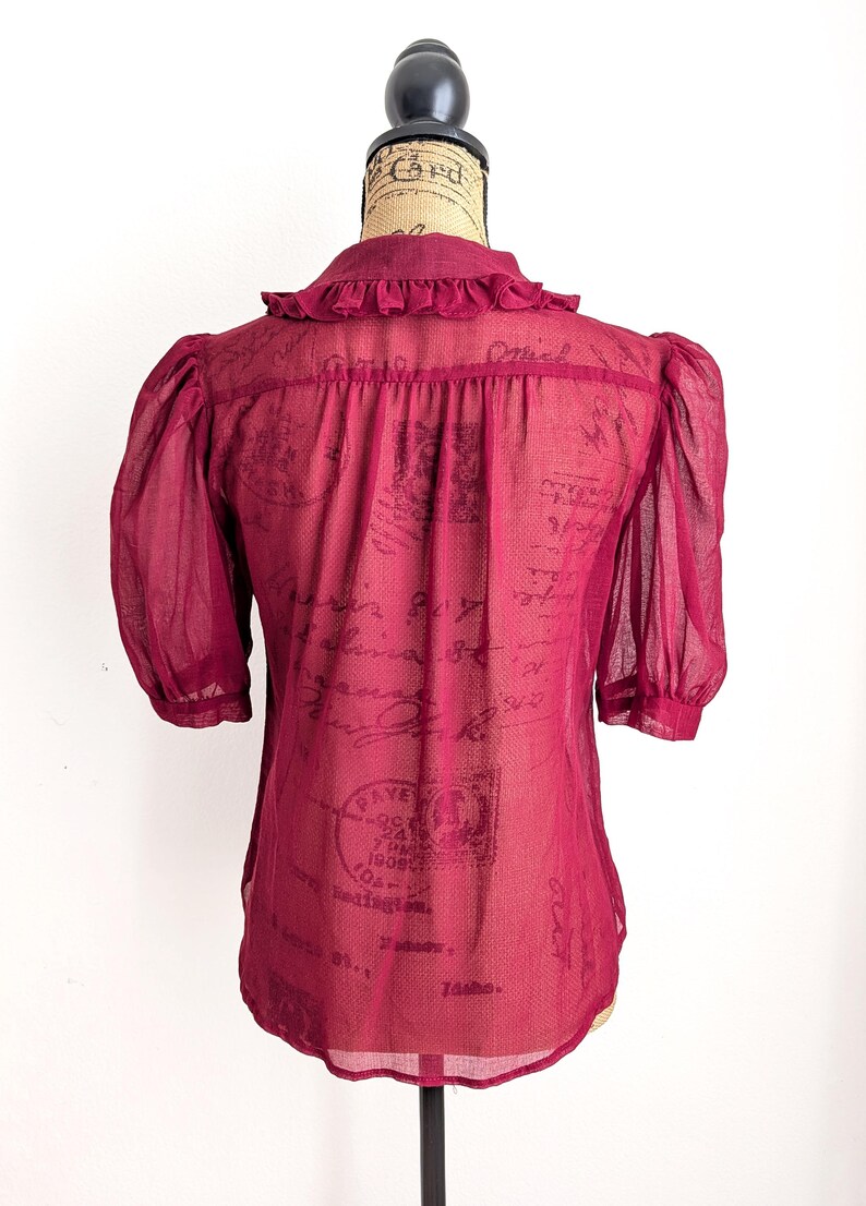 Op de afbeelding: Bordeaux transparante blouse met korte, gepofte mouwen en een kraag met ruches. De achterkant van de blouse is zichtbaar en toont een vintage printontwerp. De blouse wordt op een mannequin gepresenteerd.