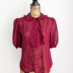 Op de afbeelding: Bordeauxkleurige, doorschijnende blouse met korte, gepofte mouwen en een gerimpelde kraag. De blouse heeft een knoopsluiting aan de voorkant met extra ruches. Het kledingstuk wordt op een mannequin getoond.
