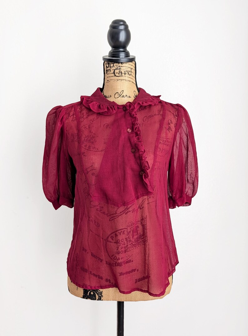 Op de afbeelding: Bordeauxkleurige, doorschijnende blouse met korte, gepofte mouwen en een gerimpelde kraag. De blouse heeft een knoopsluiting aan de voorkant met extra ruches. Het kledingstuk wordt op een mannequin getoond.