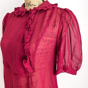 Op de afbeelding: Een doorschijnende, bordeauxrode blouse met een gerimpelde kraag en een knoopsluiting aan de voorkant. De korte mouwen zijn bij de manchetten gerimpeld. De stof heeft een subtiel patroon en een vintage esthetiek.