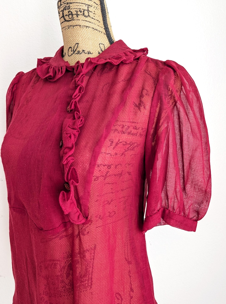 Op de afbeelding: Een doorschijnende, bordeauxrode blouse met een gerimpelde kraag en een knoopsluiting aan de voorkant. De korte mouwen zijn bij de manchetten gerimpeld. De stof heeft een subtiel patroon en een vintage esthetiek.