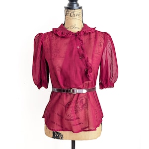Op de afbeelding: Een bordeauxrode blouse met korte, gepofte mouwen en een gerimpelde kraag. De transparante stof wordt in de taille samengeknepen met een bruine riem. De blouse heeft een knoopsluiting aan de voorkant en wordt op een mannequin gepresenteerd.