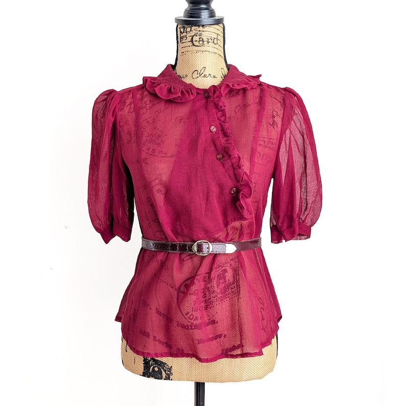 Op de afbeelding: Een bordeauxrode blouse met korte, gepofte mouwen en een gerimpelde kraag. De transparante stof wordt in de taille samengeknepen met een bruine riem. De blouse heeft een knoopsluiting aan de voorkant en wordt op een mannequin gepresenteerd.