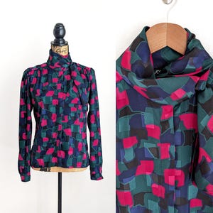 May include: A long-sleeved blouse with a high neckline and a colorful geometric pattern. The pattern features magenta, teal, navy, and black shapes. The blouse is displayed on a mannequin and a hanger.