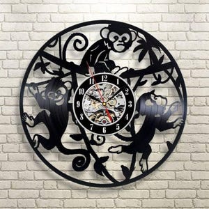 Puede incluir: Reloj de pared decorativo negro con un tema de monos. El reloj tiene una esfera redonda con un movimiento mecánico visible y manecillas negras. El diseño exterior incluye tres siluetas de monos y follaje.