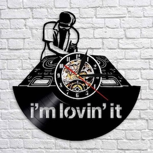 Peut inclure: Horloge en vinyle noir avec une silhouette de DJ et l'inscription "i'm lovin' it". Le cadran de l'horloge a une bordure blanche, des chiffres noirs et une trotteuse rouge. L'horloge est fixée sur un mur de briques blanches.