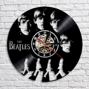 May include: Black vinyl record clock featuring the faces of The Beatles and the band's name. The clock face has a white border with black numbers and hands. The clock also features the iconic Abbey Road silhouette.